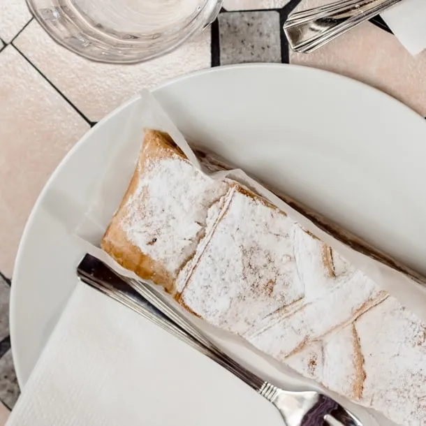 Apfelstrudel mit Staubzucker auf weißem Teller, auf einem Mosaiktisch des Cafés